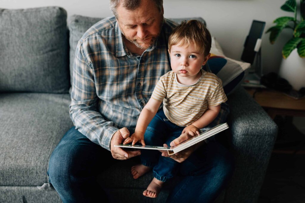 Young son looking at camera, sitting on Dad's knee while read a book, value of photography, especially plus size family photos