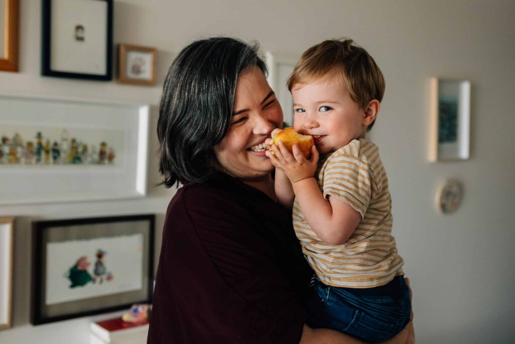 Boy eating apple, in Mum's arms, smiling at camera, plus size family photos